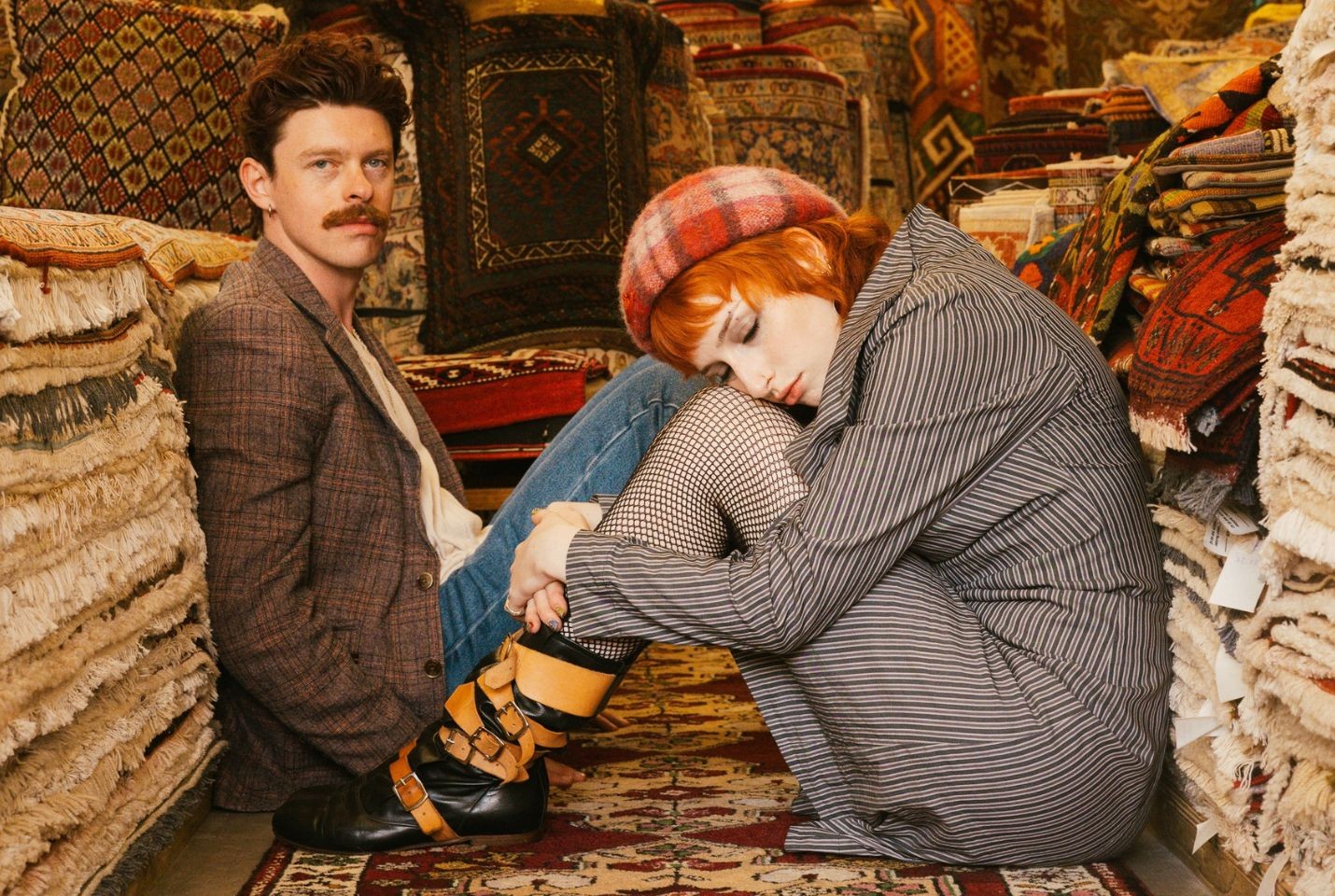 Two people sat on the floor surrounded by piles of rugs
